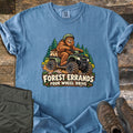 Forest Errands Four Wheel Drive Comfort Colors T-shirt in Blue Jean, laid flat. A grinning Bigfoot in a helmet rides a muddy green ATV past pine trees above a wooden sign reading Forest Errands Four Wheel Drive. Olive pants and brown hiking boots lie beside it on weathered wood.