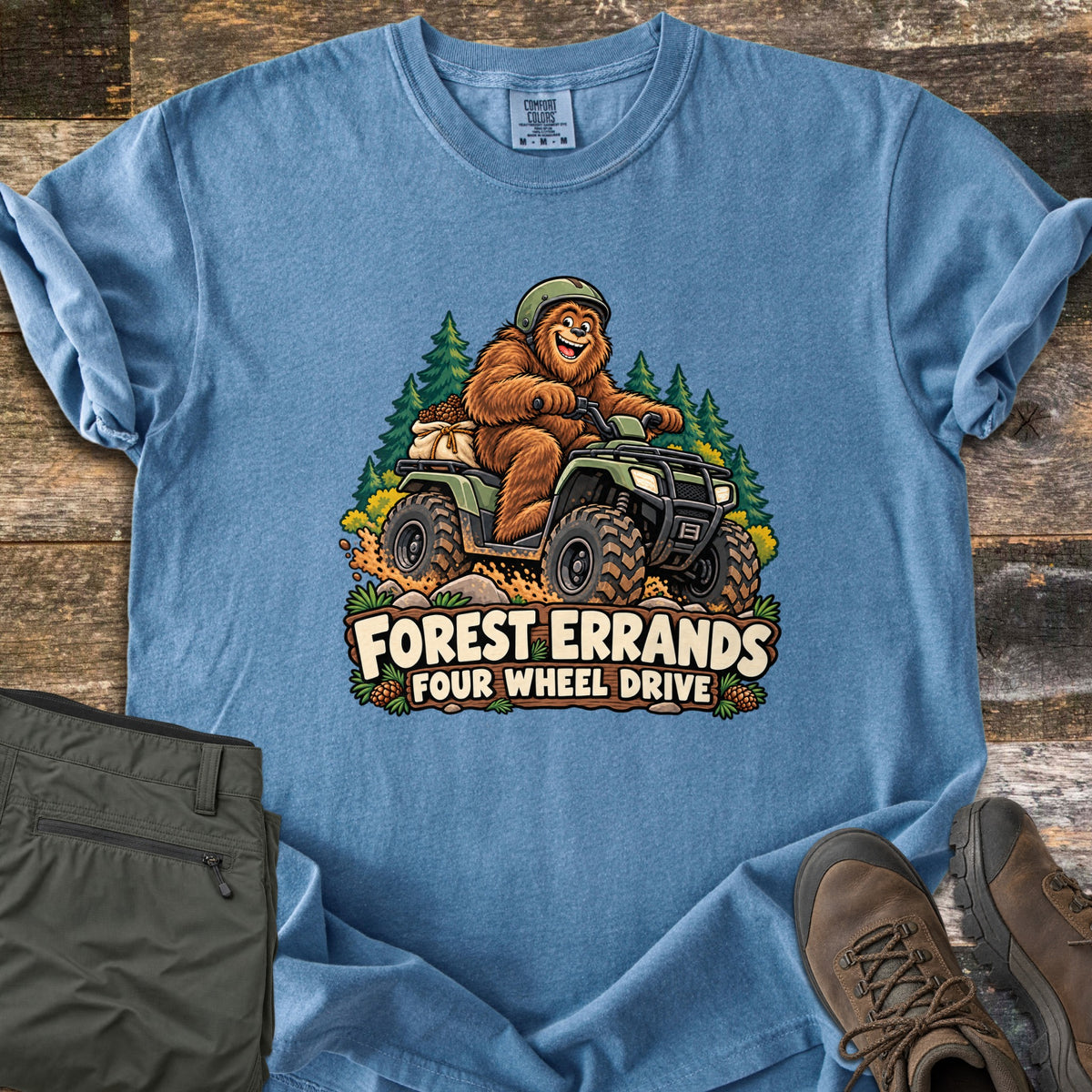 Forest Errands Four Wheel Drive Comfort Colors T-shirt in Blue Jean, laid flat. A grinning Bigfoot in a helmet rides a muddy green ATV past pine trees above a wooden sign reading Forest Errands Four Wheel Drive. Olive pants and brown hiking boots lie beside it on weathered wood.