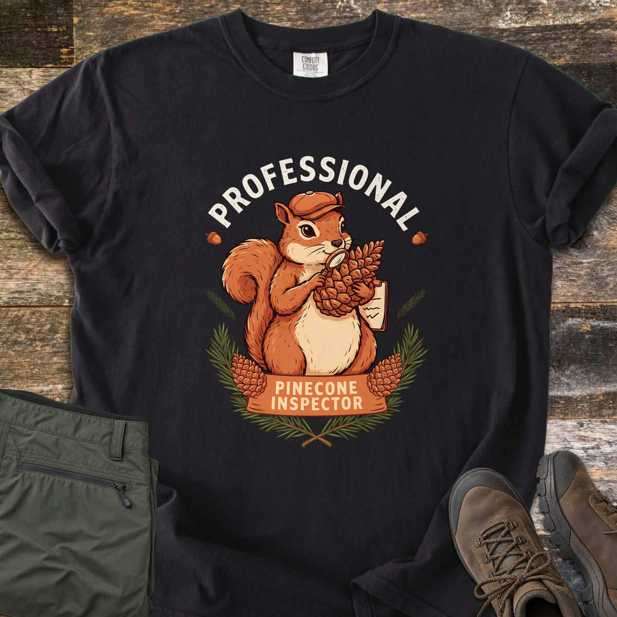 Professional Pinecone Inspector Comfort Colors T-shirt in Black, laid flat. A cartoon squirrel in an orange cap studies a pinecone with a magnifying glass, holding a clipboard, under the word Professional, with Pinecone Inspector on a banner. Hiking pants and boots lie on wood.