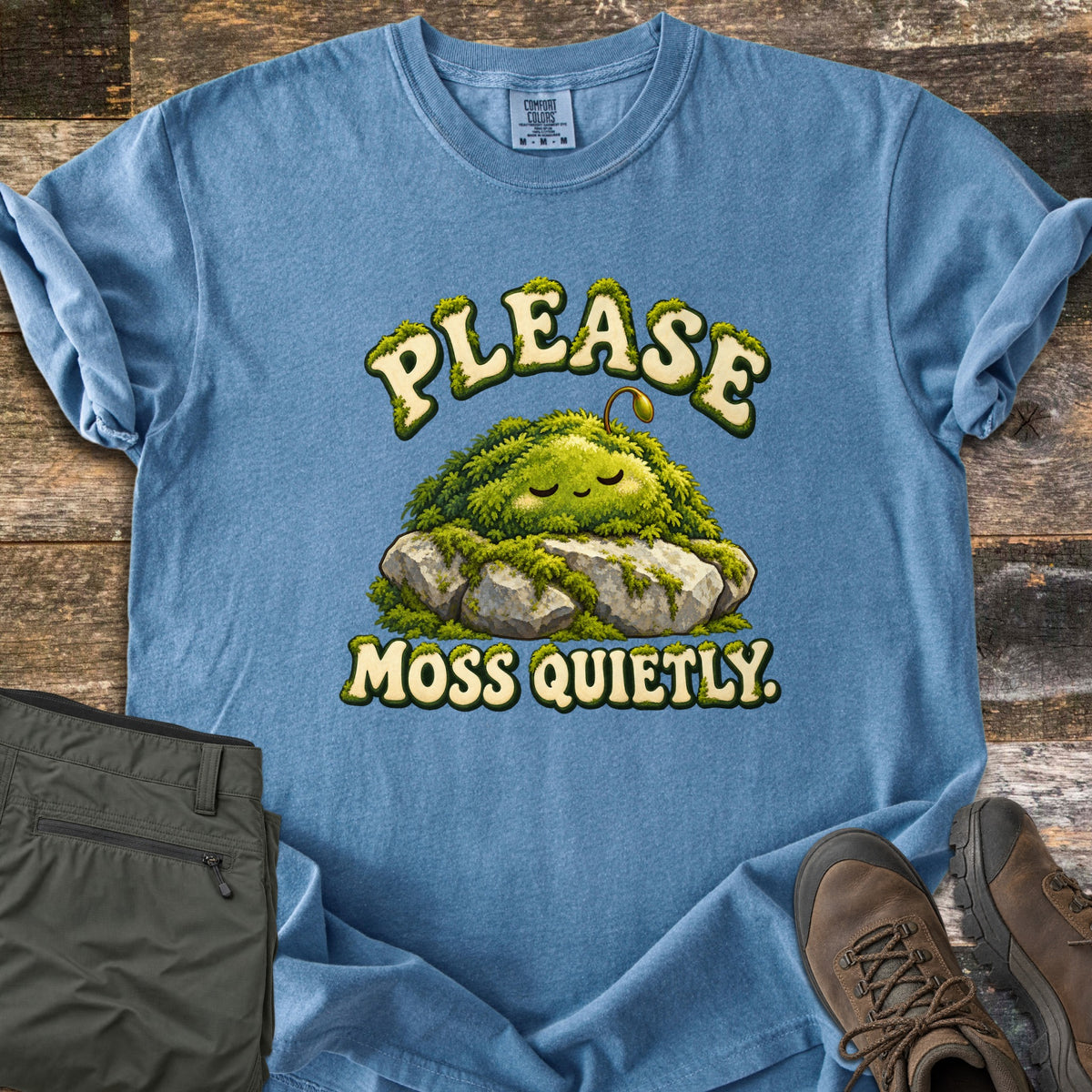 Please Moss Quietly Comfort Colors T-shirt in Blue Jean, laid flat. A sleepy, smiling clump of moss with a tiny sprout rests on gray rocks, with PLEASE arched above and MOSS QUIETLY. below in moss-covered letters. Olive hiking pants and brown hiking boots lie on weathered wood.
