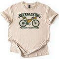 Bikepacking Packed for Detours Comfort Colors T-shirt in Sand, laid flat. A dark green mountain bike with mustard yellow frame, saddle and handlebar bags sits between the words BIKEPACKING and PACKED FOR DETOURS. The shirt lies on a white surface with cuffed sleeves.