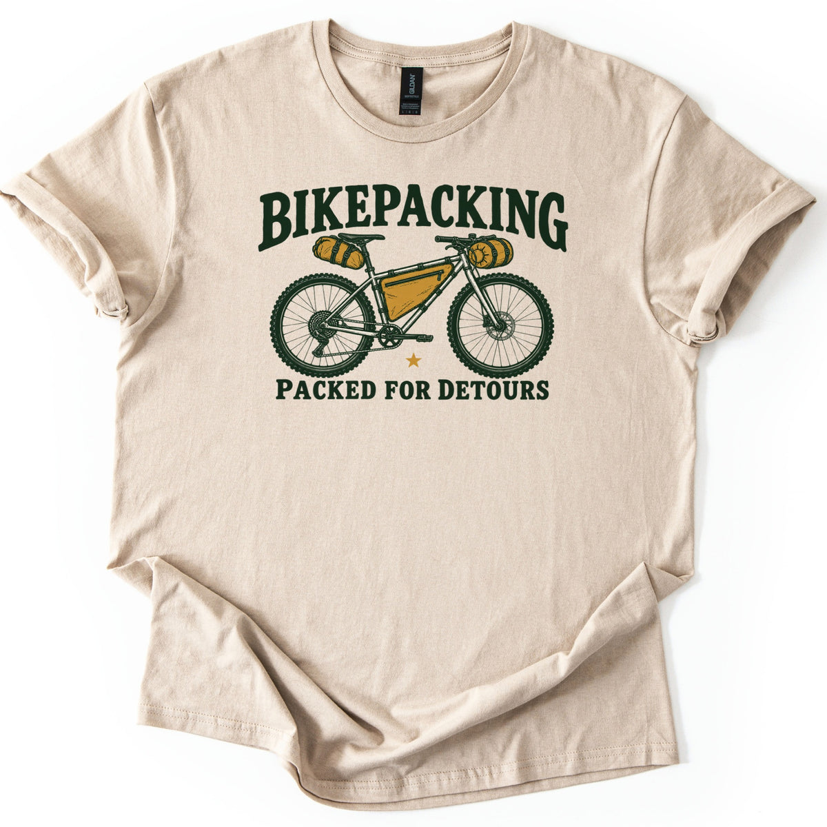 Bikepacking Packed for Detours Comfort Colors T-shirt in Sand, laid flat. A dark green mountain bike with mustard yellow frame, saddle and handlebar bags sits between the words BIKEPACKING and PACKED FOR DETOURS. The shirt lies on a white surface with cuffed sleeves.
