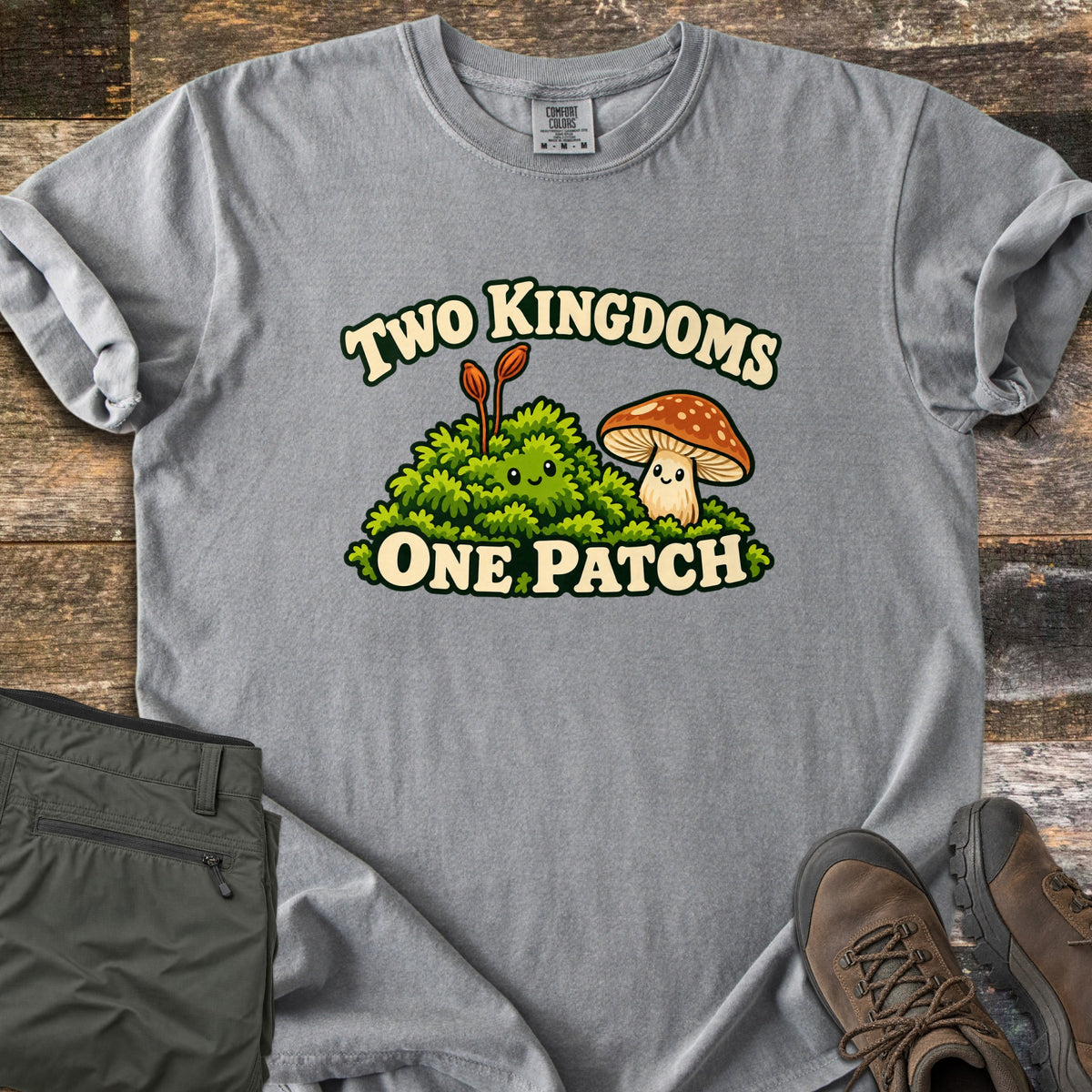 Two Kingdoms One Patch Comfort Colors T-shirt in Grey, laid flat. A smiling moss clump with two orange stalks sits beside a smiling spotted brown mushroom, between the words Two Kingdoms and One Patch. Sleeves rolled, on weathered wood with olive hiking pants and brown boots.