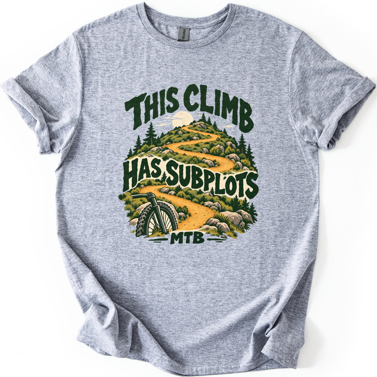 This Climb Has Subplots Comfort Colors T-shirt in Sport Grey, laid flat. A winding dirt trail climbs a rocky, pine-covered hill toward the sun, with a mountain bike's front wheel at the bottom, framed by the words This Climb Has Subplots and MTB in dark green. On a white background.