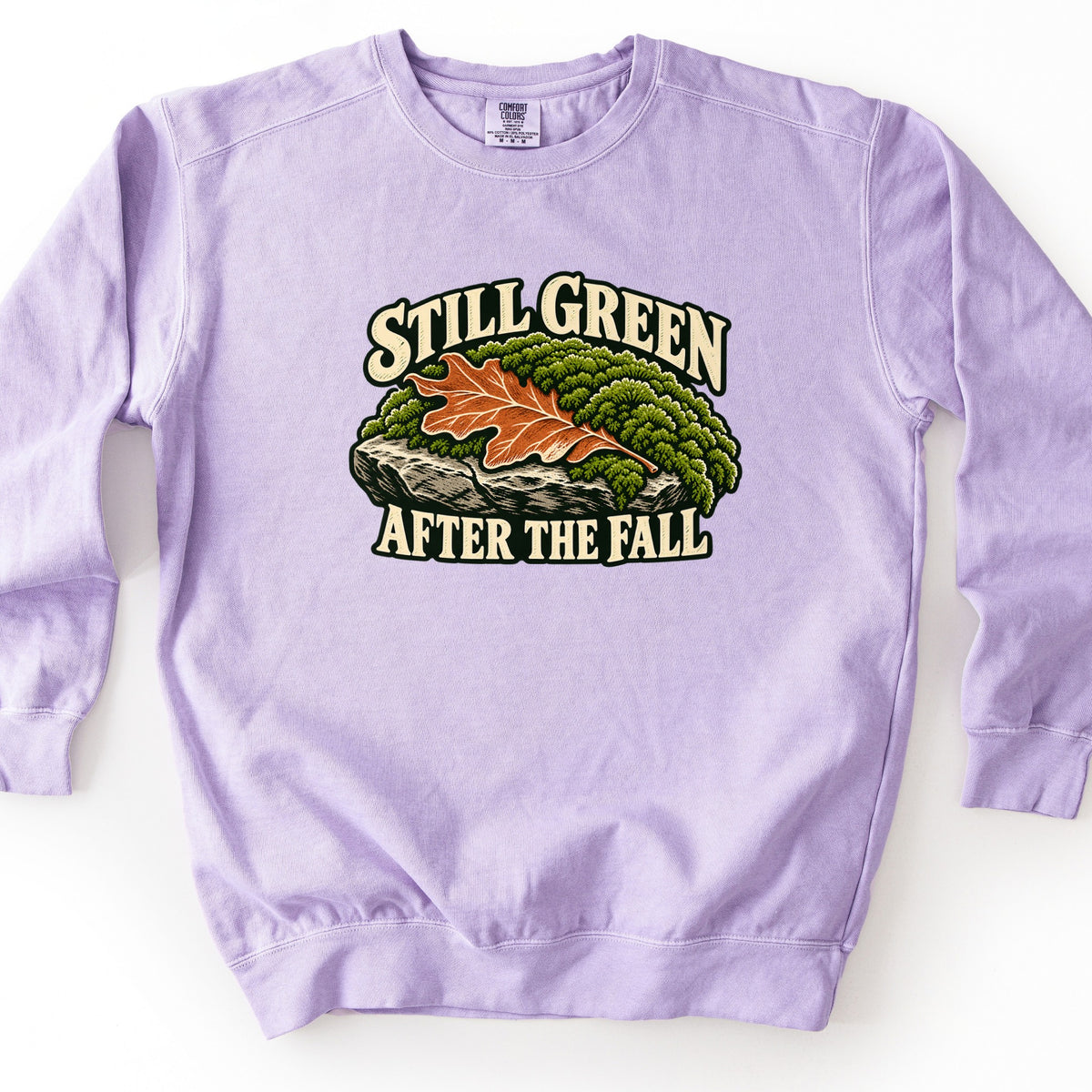 Still Green After the Fall Comfort Colors crewneck sweatshirt in Orchid, laid flat. An orange oak leaf rests on green moss covering a gray rock, with the words Still Green arched above and After the Fall below in cream letters outlined in dark green. It lies on a white surface.