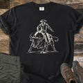 Cowboy Bull Rider Comfort Colors T-shirt in Black, laid flat. A cream line drawing of a cowboy in a hat and vest riding a charging longhorn bull, no words. The shirt lies on weathered wood with folded olive pants and brown hiking boots.
