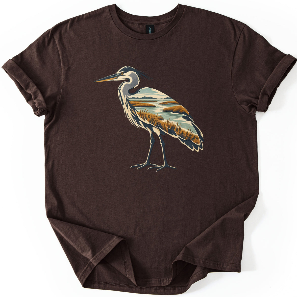 Heron Marsh Comfort Colors T-shirt in Dark Chocolate, laid flat. A standing heron in profile, its body filled with a marsh scene of winding water, golden grasses and distant hills. No words are printed. The sleeves are cuffed, on a white background.