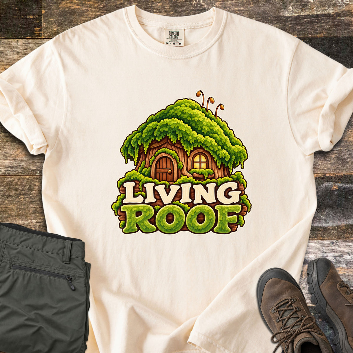 Living Roof Comfort Colors T-shirt in Ivory, laid flat. A cartoon tree-stump cottage with a wooden door, glowing window, and thick green moss roof sits above the words Living Roof in cream and mossy green letters. Olive hiking pants and brown hiking boots lie on weathered wood.