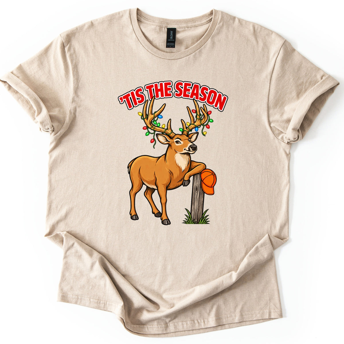 Buck Tis The Season Comfort Colors T-shirt in Sand, laid flat. Red arched text reads 'TIS THE SEASON above a cartoon buck with colored Christmas lights strung on its antlers, leaning on a wooden post that holds an orange hunting cap. Sleeves rolled, on a white surface.
