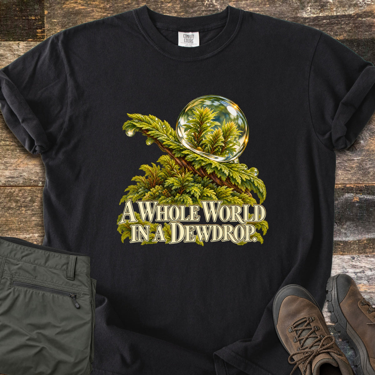 A Whole World in a Dewdrop Comfort Colors T-shirt in Black, laid flat. A clear dewdrop reflecting tiny ferns rests on a mossy green branch above the words A Whole World in a Dewdrop. It lies on weathered wood beside folded olive hiking pants and brown hiking boots.
