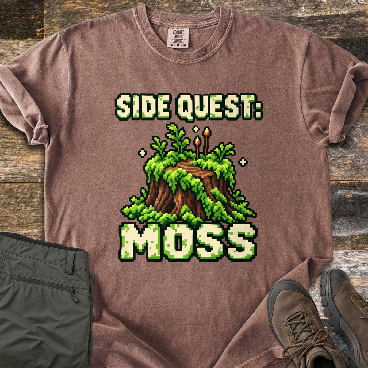 Side Quest Moss Comfort Colors T-shirt in Espresso, laid flat. Pixel art of a moss-covered tree stump with ferns, two small sporophytes and sparkles, between the words Side Quest: and Moss. Olive cargo pants and brown hiking boots lie beside it on weathered wood planks.