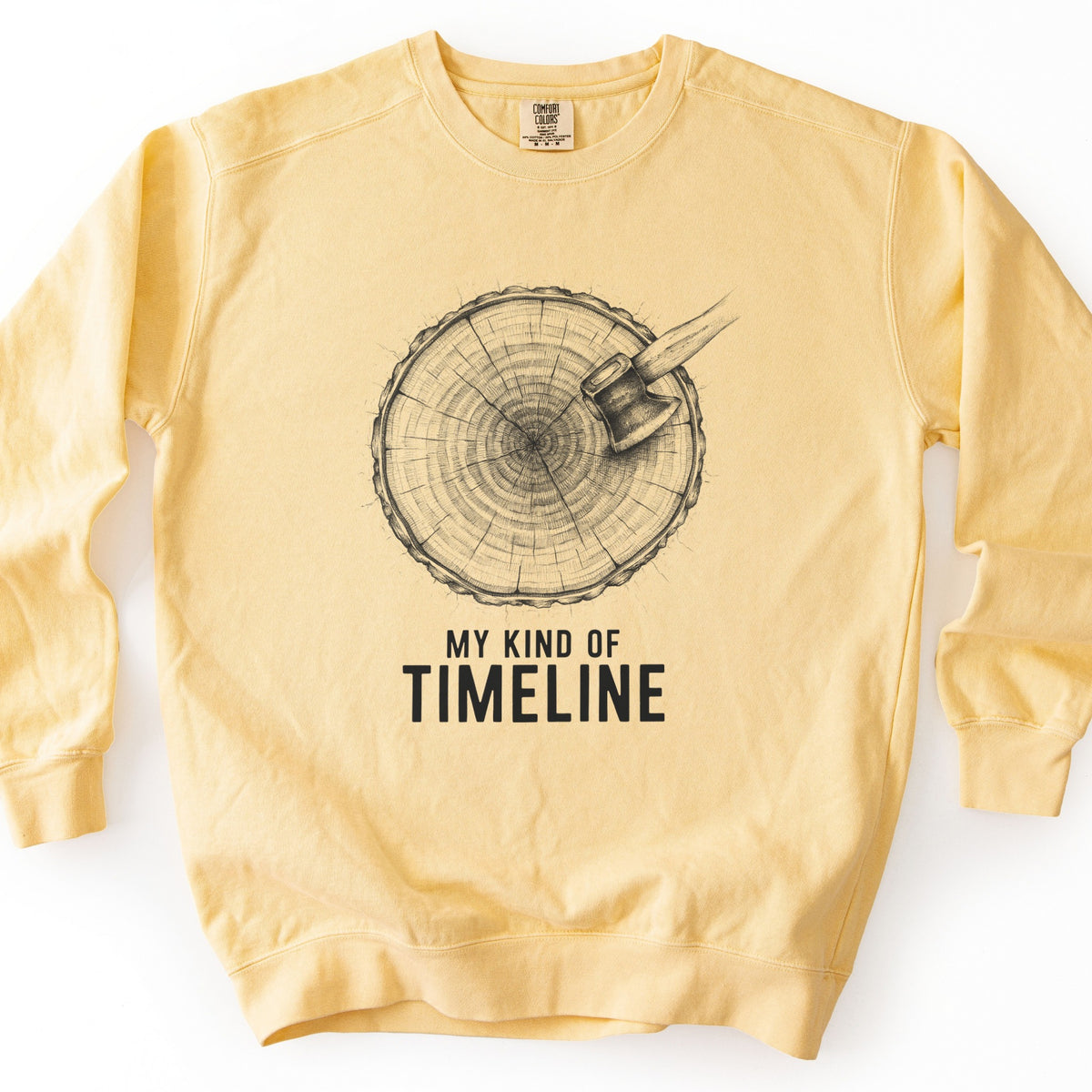 My Kind Of Timeline Comfort Colors crewneck sweatshirt in Butter, laid flat. A detailed black sketch of a cut tree stump showing its growth rings, with an axe stuck in the top right edge, above the words My Kind Of Timeline. The sweatshirt lies on a white background.