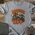 Logging Off Comfort Colors T-shirt in Grey, laid flat. A cartoon beaver in a helmet rides a muddy ATV hauling a log, between the words Logging Off. Folded olive pants and brown hiking boots sit beside the shirt on a weathered wood surface.