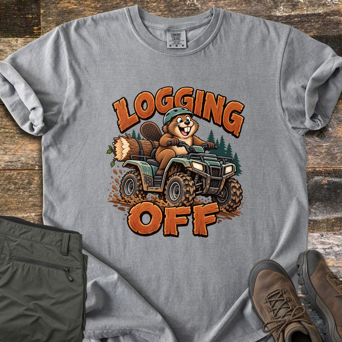Logging Off Comfort Colors T-shirt in Grey, laid flat. A cartoon beaver in a helmet rides a muddy ATV hauling a log, between the words Logging Off. Folded olive pants and brown hiking boots sit beside the shirt on a weathered wood surface.