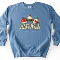 Waiting Is a Water Sport Comfort Colors crewneck sweatshirt in Blue Jean, laid flat. Three vintage fishing bobbers float in rippling water: one red and white, one cream and green, one cream with a red stripe. Below them, cream lettering reads Waiting Is a Water Sport. Set on a white background.