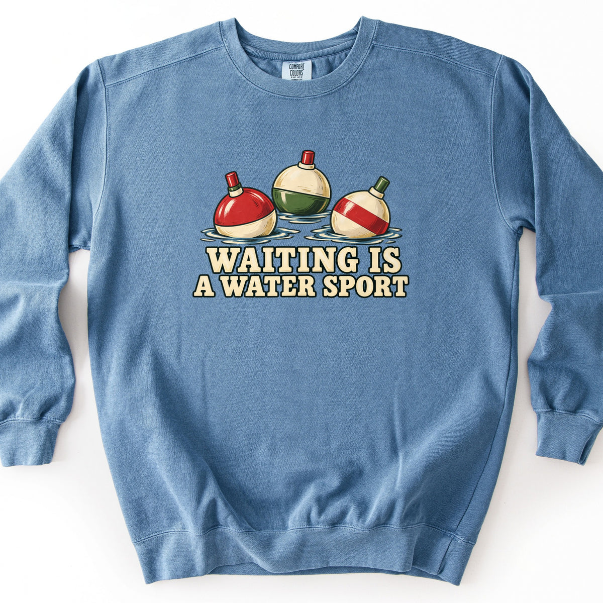Waiting Is a Water Sport Comfort Colors crewneck sweatshirt in Blue Jean, laid flat. Three vintage fishing bobbers float in rippling water: one red and white, one cream and green, one cream with a red stripe. Below them, cream lettering reads Waiting Is a Water Sport. Set on a white background.