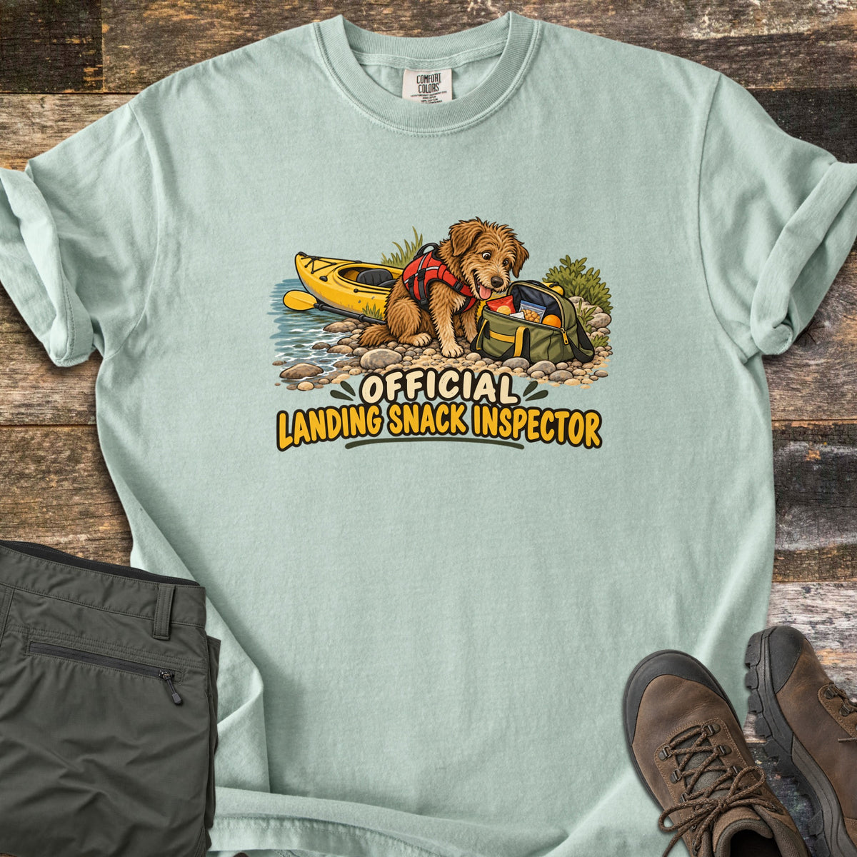Landing Snack Inspector Comfort Colors T-shirt in Bay, laid flat. A shaggy brown dog in a red life vest digs into an open green snack bag on a rocky shore beside a yellow kayak, above the words Official Landing Snack Inspector. Gray pants and brown hiking boots lie on wood planks.