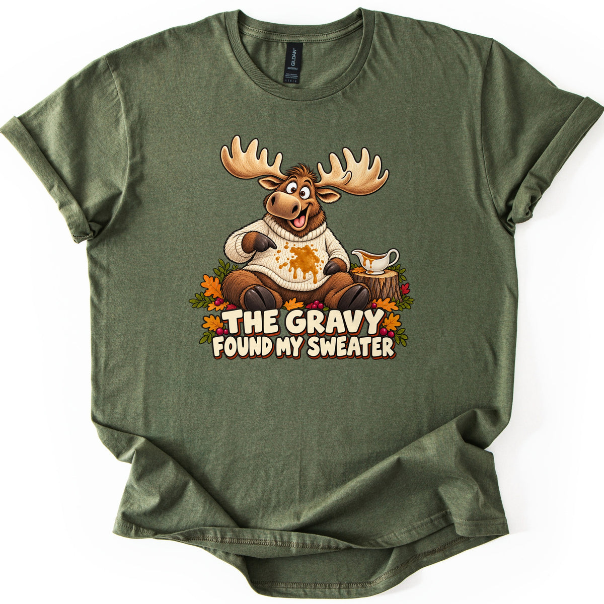 The Gravy Found My Sweater Comfort Colors T-shirt in Military Green, laid flat. A cartoon moose in a cream knit sweater points at a gravy stain, beside a gravy boat on a tree stump and autumn leaves, above the words The Gravy Found My Sweater. Sleeves are rolled on a white background.