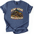 Low Gear High Expectations Comfort Colors T-shirt in Heather Navy, laid flat. A helmeted rider drives a mud-splattered yellow ATV over a grassy dirt mound between the words Low Gear and High Expectations in cream brush lettering. Sleeves are cuffed on a white background.