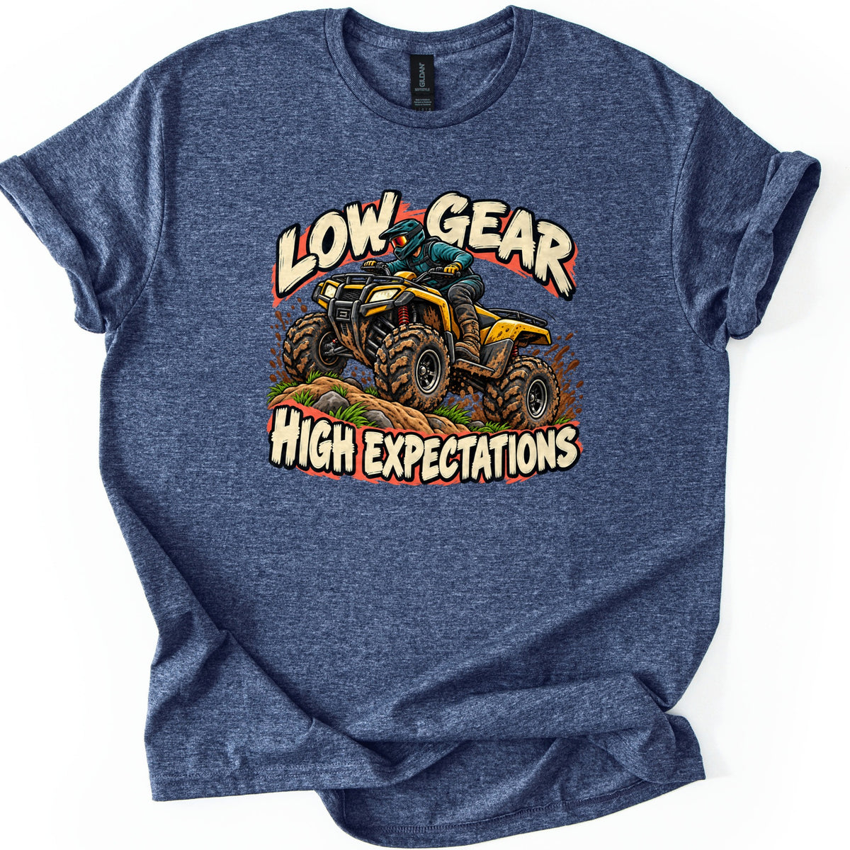 Low Gear High Expectations Comfort Colors T-shirt in Heather Navy, laid flat. A helmeted rider drives a mud-splattered yellow ATV over a grassy dirt mound between the words Low Gear and High Expectations in cream brush lettering. Sleeves are cuffed on a white background.