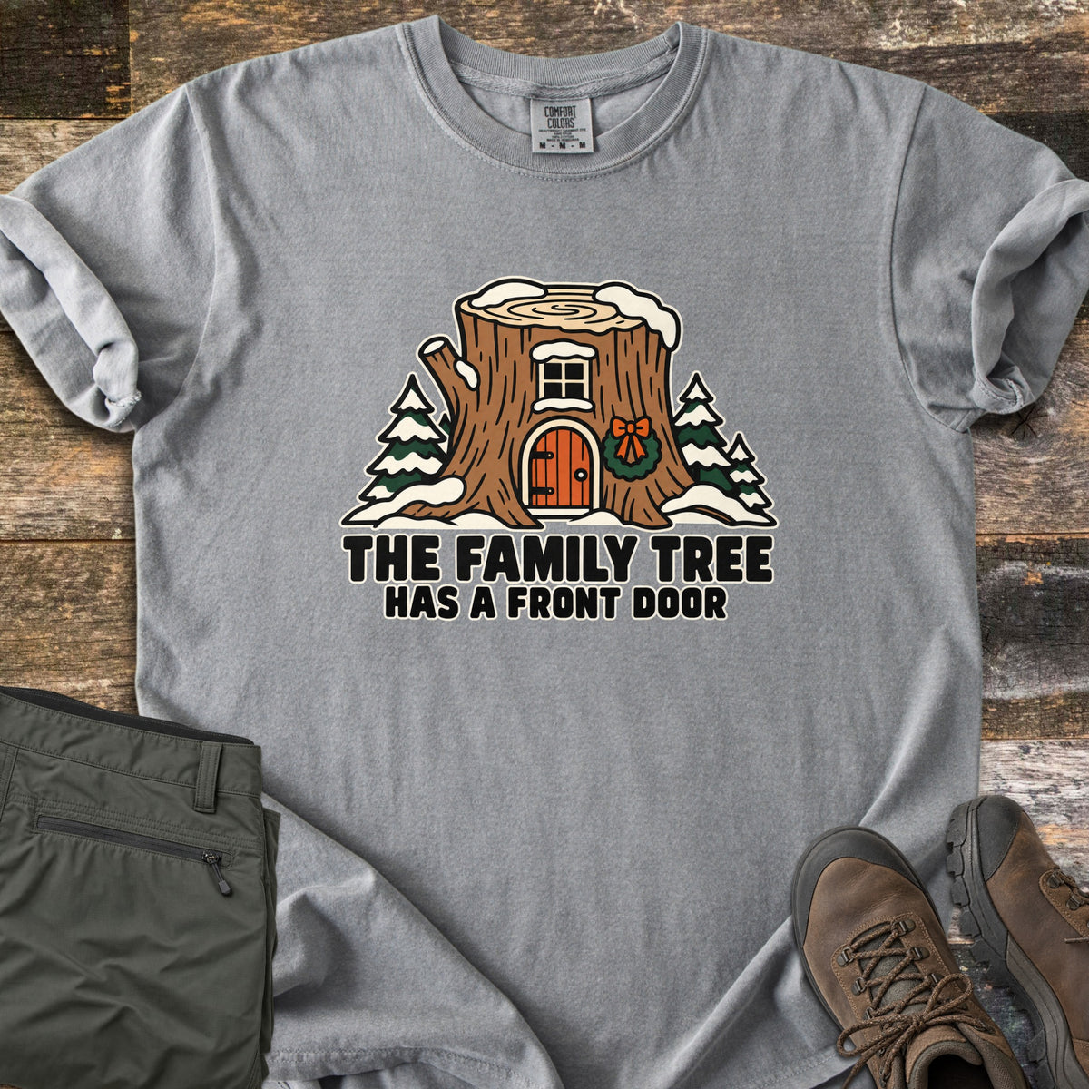The Family Tree Has a Front Door Comfort Colors T-shirt in Grey, laid flat. A snowy tree stump house with a window, an orange arched door, and a wreath with a bow sits between pine trees above the words The Family Tree Has a Front Door. Olive pants and brown hiking boots lie on dark wood.