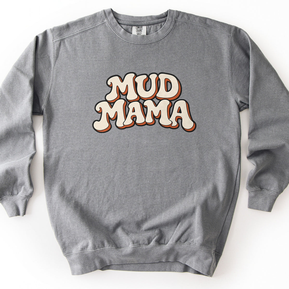 Mud Mama Comfort Colors crewneck sweatshirt in Grey, laid flat. The words Mud Mama are printed across the chest in large bubbly retro letters, cream with a black outline and a rust orange drop shadow. The sweatshirt lies on a plain white surface.