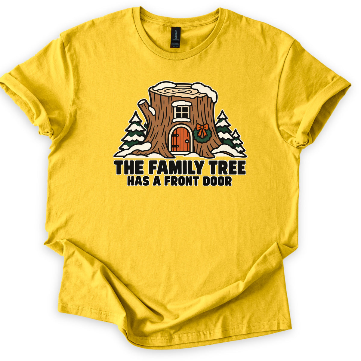 The Family Tree Has a Front Door Comfort Colors T-shirt in Daisy, laid flat. A snow-topped tree stump with a small window, an arched orange door and a wreath with a bow sits between snowy pine trees above the words The Family Tree Has a Front Door. Cuffed sleeves, white background.
