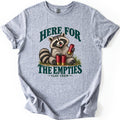 Here for the Empties Comfort Colors T-shirt in Sport Grey, laid flat. A grinning raccoon sits in grass holding a red shotgun shell, with two more at its feet, between the words Here For The Empties and Clay Crew in dark green. Sleeves are cuffed, on a white background.