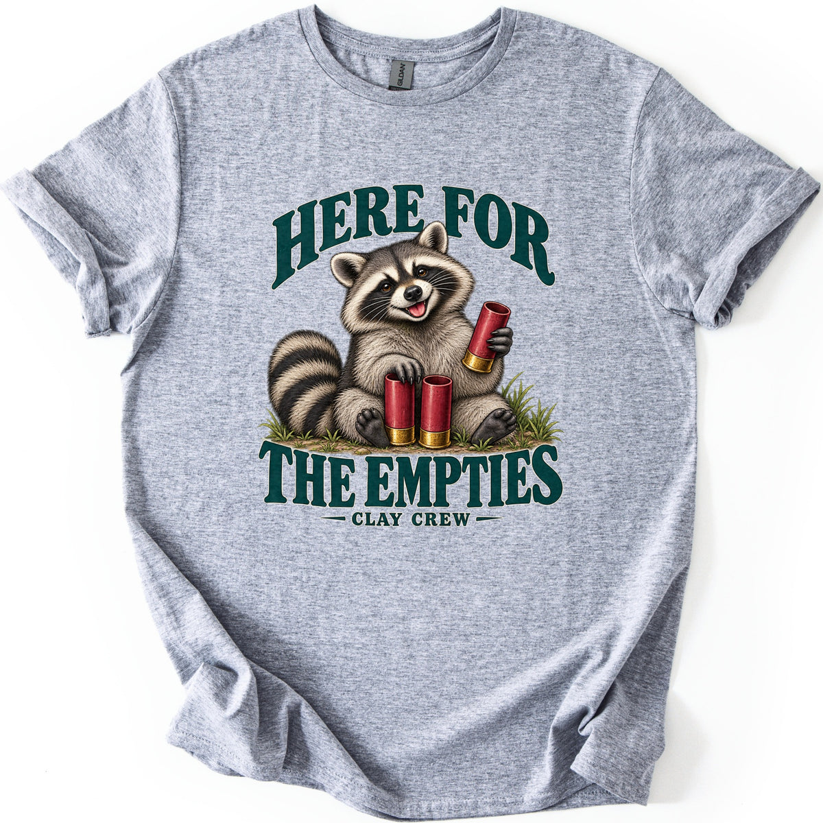 Here for the Empties Comfort Colors T-shirt in Sport Grey, laid flat. A grinning raccoon sits in grass holding a red shotgun shell, with two more at its feet, between the words Here For The Empties and Clay Crew in dark green. Sleeves are cuffed, on a white background.