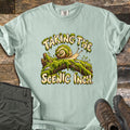 Taking the Scenic Inch Comfort Colors T-shirt in Bay, laid flat. A smiling cartoon snail with a spiral shell crawls along a mossy log between the words Taking the Scenic Inch., lettered in cream with dripping moss. Olive hiking pants and brown boots sit on a weathered wood floor.