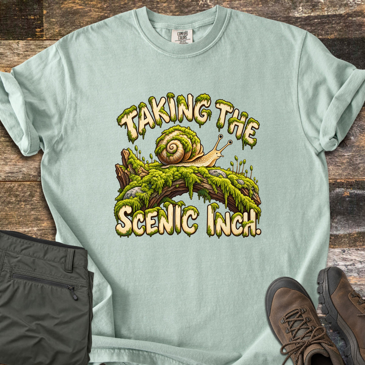 Taking the Scenic Inch Comfort Colors T-shirt in Bay, laid flat. A smiling cartoon snail with a spiral shell crawls along a mossy log between the words Taking the Scenic Inch., lettered in cream with dripping moss. Olive hiking pants and brown boots sit on a weathered wood floor.