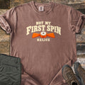 Not My First Spin Comfort Colors T-shirt in Espresso, laid flat. Cream arched text reads Not My First Spin above an orange two-winged helice target with a white center, with Helice below. Folded olive pants and brown hiking boots sit beside it on weathered wood planks.