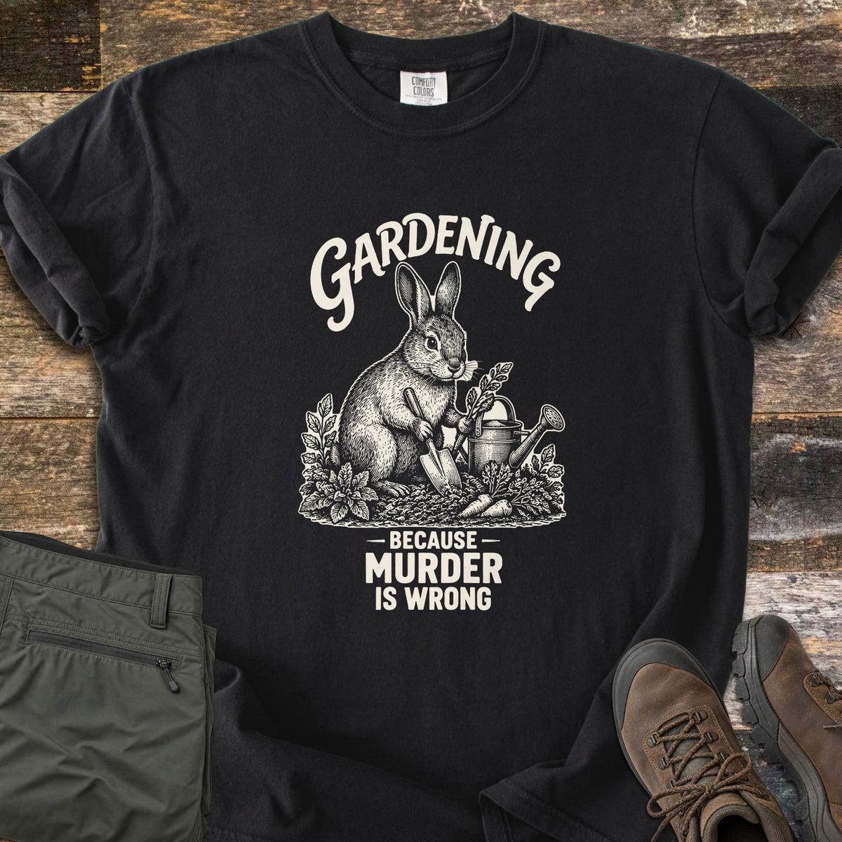 Gardening Murder Rabbit Comfort Colors T-shirt in Black, laid flat. An etched-style rabbit holds a trowel beside a watering can, carrots and plants, under the word Gardening and above Because Murder Is Wrong. Green pants and brown hiking boots sit on weathered wood.