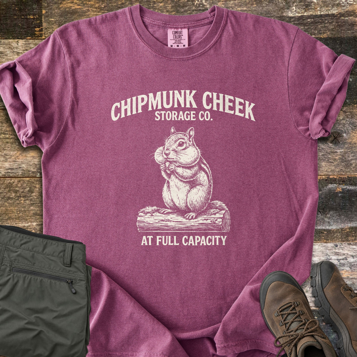 Chipmunk Cheek Storage Co Comfort Colors T-shirt in Berry, laid flat. Cream print of a chipmunk with full cheeks sitting on a log, under the words Chipmunk Cheek Storage Co. and above At Full Capacity. Rolled sleeves, on weathered wood with olive pants and brown hiking boots.