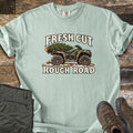 Fresh Cut Rough Road Comfort Colors T-shirt in Bay, laid flat. An army-green ATV with muddy tires carries a strapped-on pine tree over snowy ground, between the words Fresh Cut and Rough Road. Shown on wood planks with olive cargo pants and brown hiking boots.