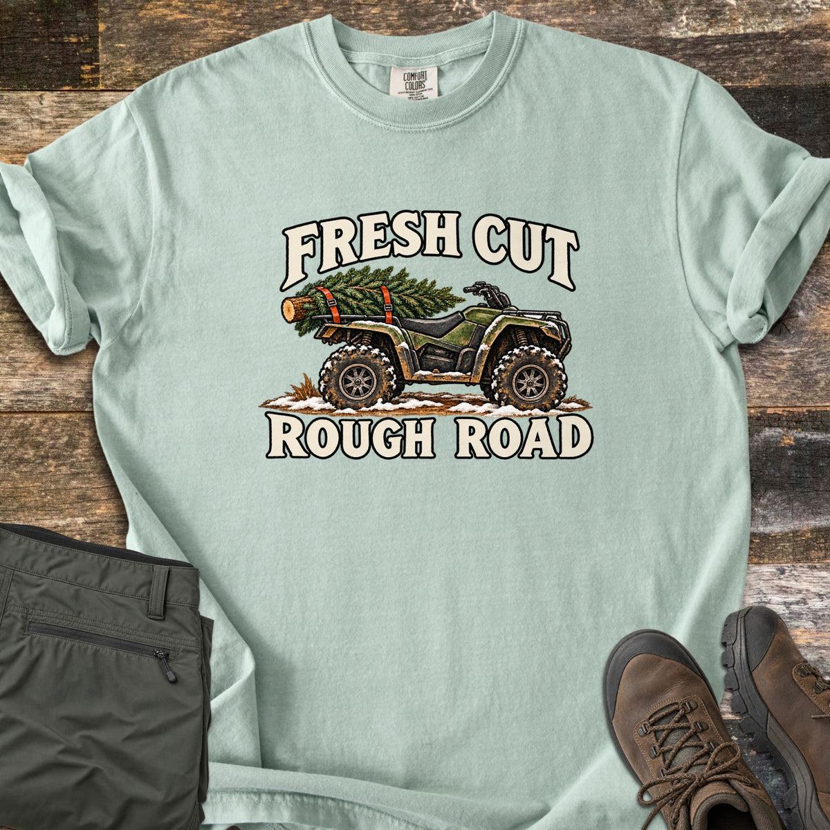 Fresh Cut Rough Road Comfort Colors T-shirt in Bay, laid flat. An army-green ATV with muddy tires carries a strapped-on pine tree over snowy ground, between the words Fresh Cut and Rough Road. Shown on wood planks with olive cargo pants and brown hiking boots.