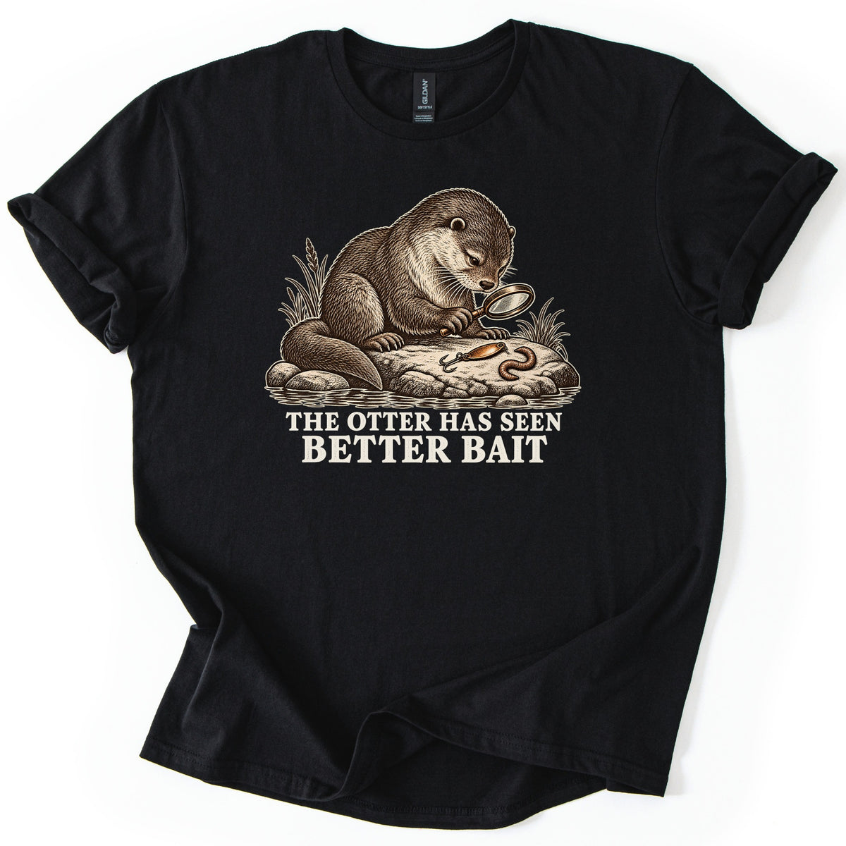 The Otter Has Seen Better Bait Comfort Colors T-shirt in Black, laid flat. An illustrated otter on a riverside rock inspects a fishing lure and a worm through a magnifying glass, above the words The Otter Has Seen Better Bait. The sleeves are rolled on a white background.