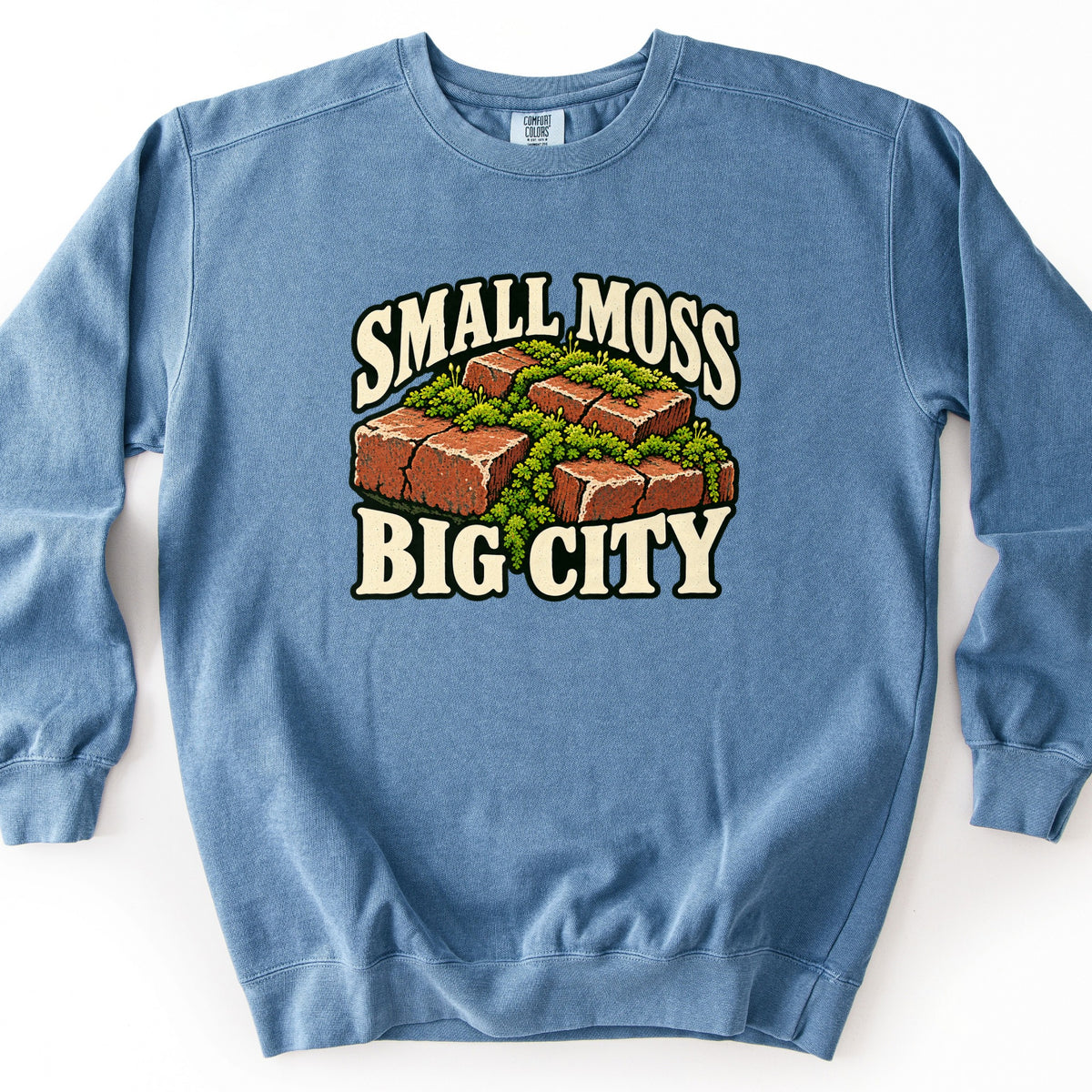 Small Moss Big City Comfort Colors crewneck sweatshirt in Blue Jean, laid flat. Front print shows red bricks with bright green moss growing between them, framed by cream arched lettering reading Small Moss above and Big City below. Shown on a white background.