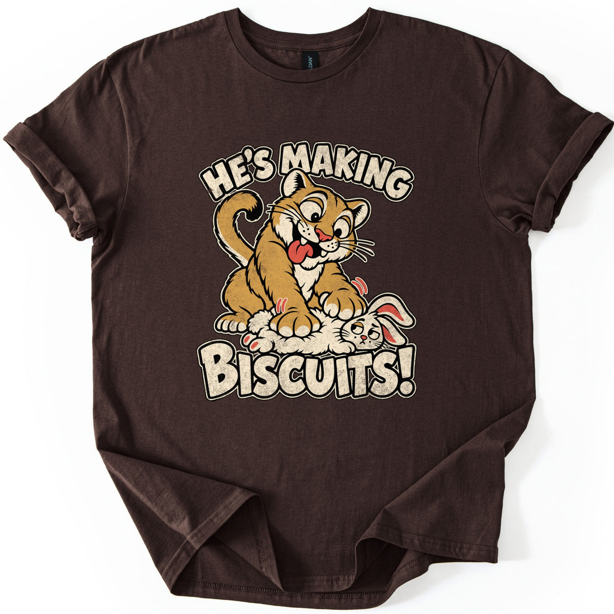 He's Making Biscuits Comfort Colors T-shirt in Dark Chocolate, laid flat. A grinning cartoon cougar with its tongue out kneads a dazed white rabbit on its back, with the words He's Making Biscuits! in distressed cream letters. The shirt lies on a white background with cuffed sleeves.