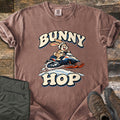 Bunny Hop Comfort Colors T-shirt in Espresso, laid flat. A cartoon bunny in a white helmet rides a navy, white and orange snowmobile over a snowbank between the words Bunny Hop. Folded green pants and brown hiking boots lie beside it on a wood floor.