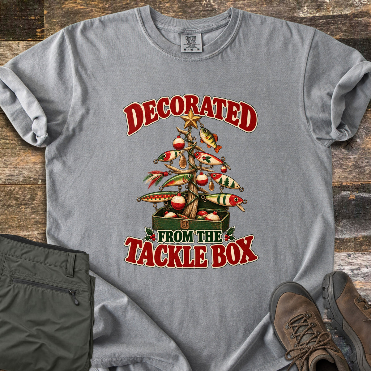 Decorated From The Tackle Box Comfort Colors T-shirt in Grey, laid flat. A Christmas tree made of driftwood is hung with fishing lures, bobbers and a star, rising from a green tackle box, between the words Decorated From The Tackle Box. Olive pants and brown boots sit on wood planks.