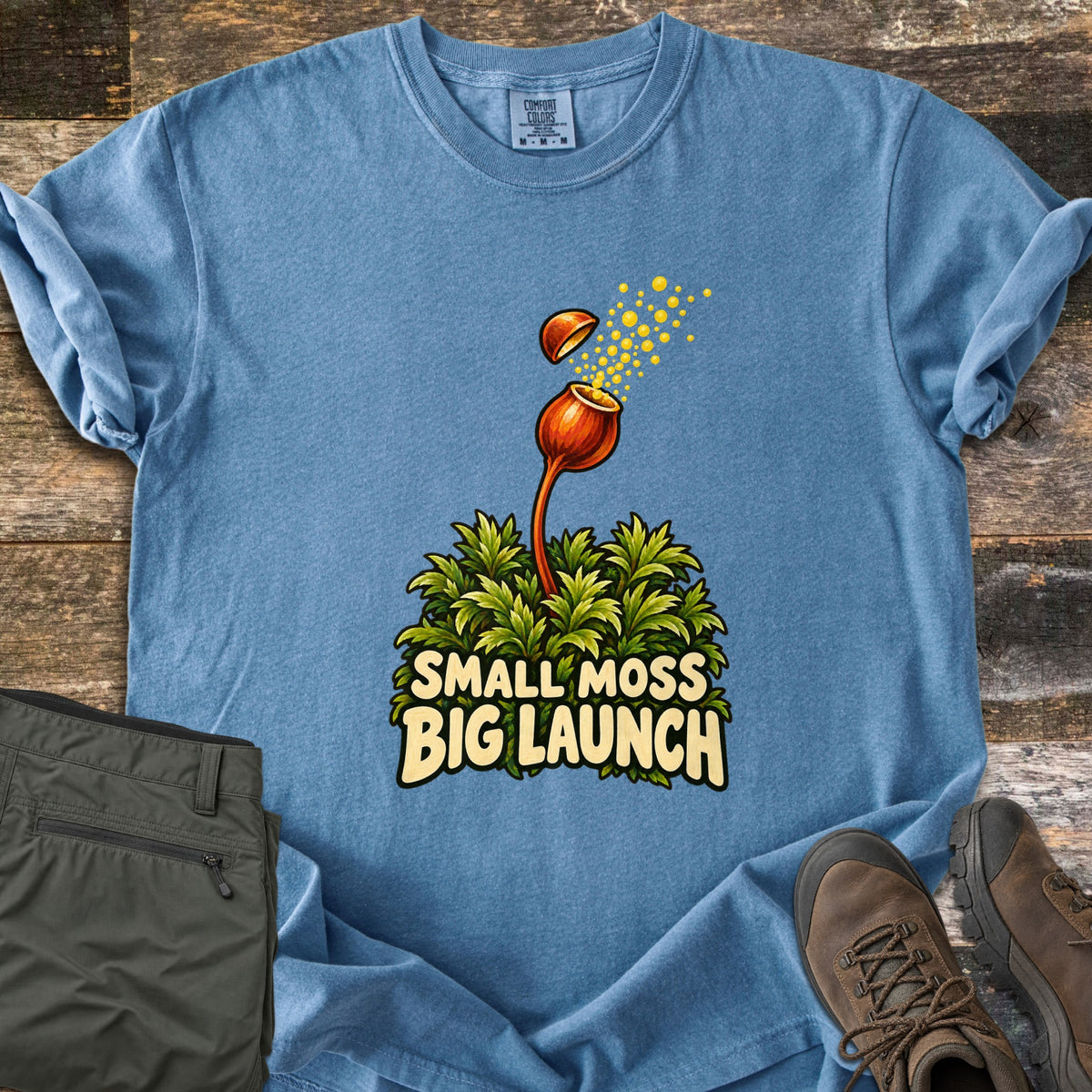 Small Moss Big Launch Comfort Colors T-shirt in Blue Jean, laid flat. An orange moss capsule on a tall stalk pops its lid and bursts yellow spores above a green moss clump and the words Small Moss Big Launch. Olive hiking pants and brown hiking boots sit beside it on weathered wood.