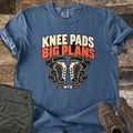 Knee Pads Big Plans Comfort Colors T-shirt in Denim, laid flat. The words Knee Pads Big Plans in cream and orange sit above a pair of black and cream mountain bike knee pads, with MTB below. Olive cargo pants and brown hiking boots lie beside it on weathered wood.