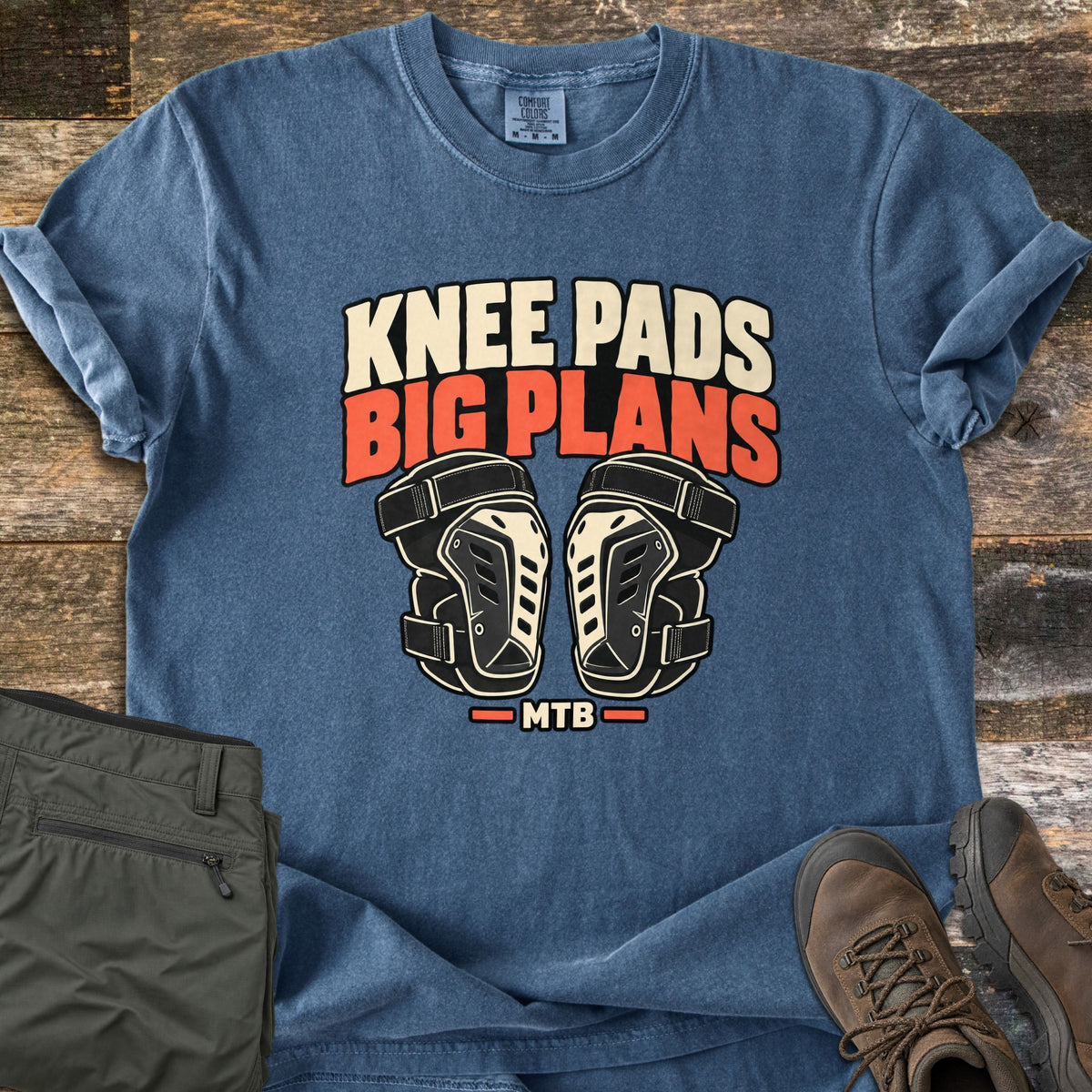 Knee Pads Big Plans Comfort Colors T-shirt in Denim, laid flat. The words Knee Pads Big Plans in cream and orange sit above a pair of black and cream mountain bike knee pads, with MTB below. Olive cargo pants and brown hiking boots lie beside it on weathered wood.