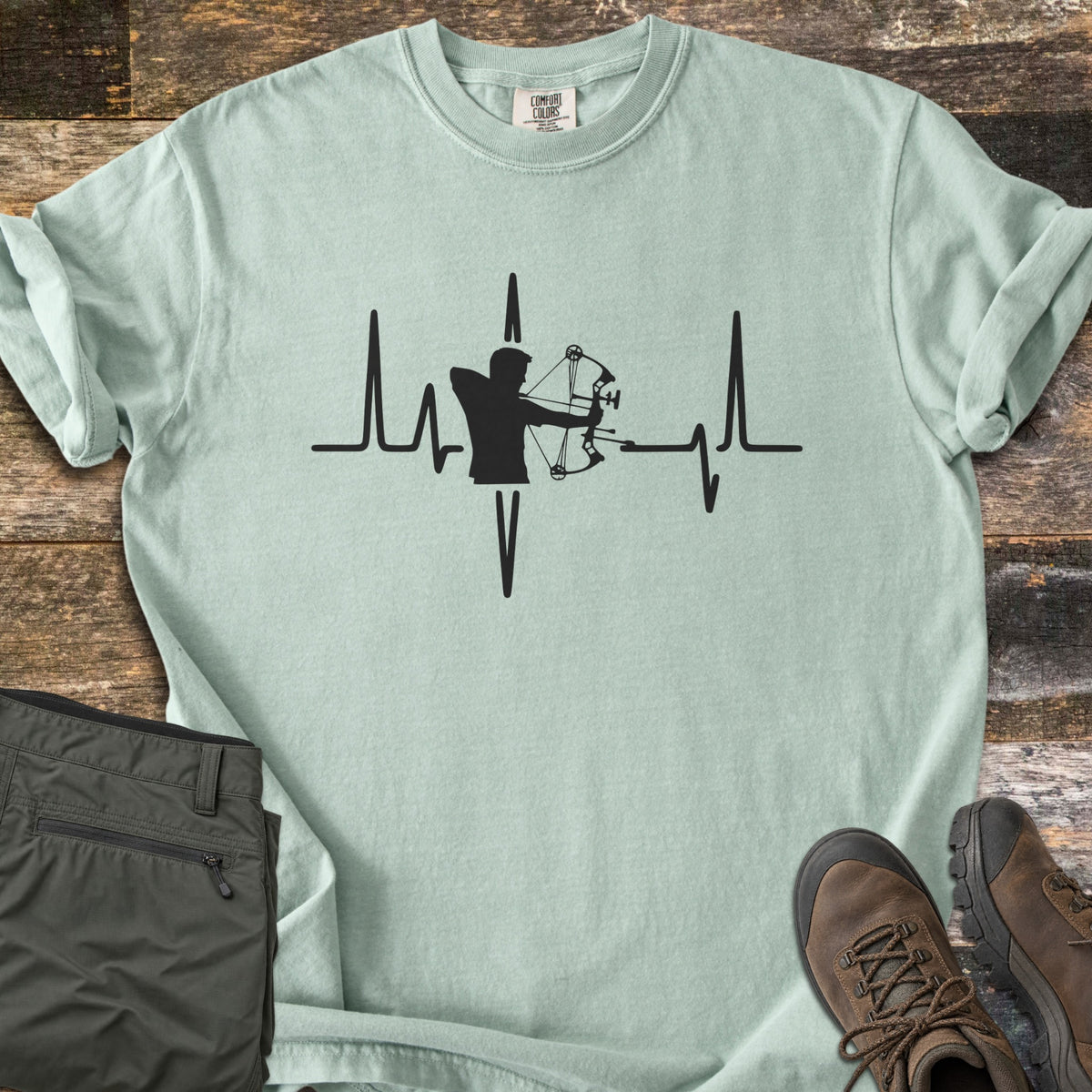Compound Bow Heartbeat Comfort Colors T-shirt in Bay, laid flat. A black heartbeat line with the silhouette of an archer drawing a compound bow at its center. Sleeves are rolled on a wood plank floor, with olive cargo pants and brown hiking boots beside it.
