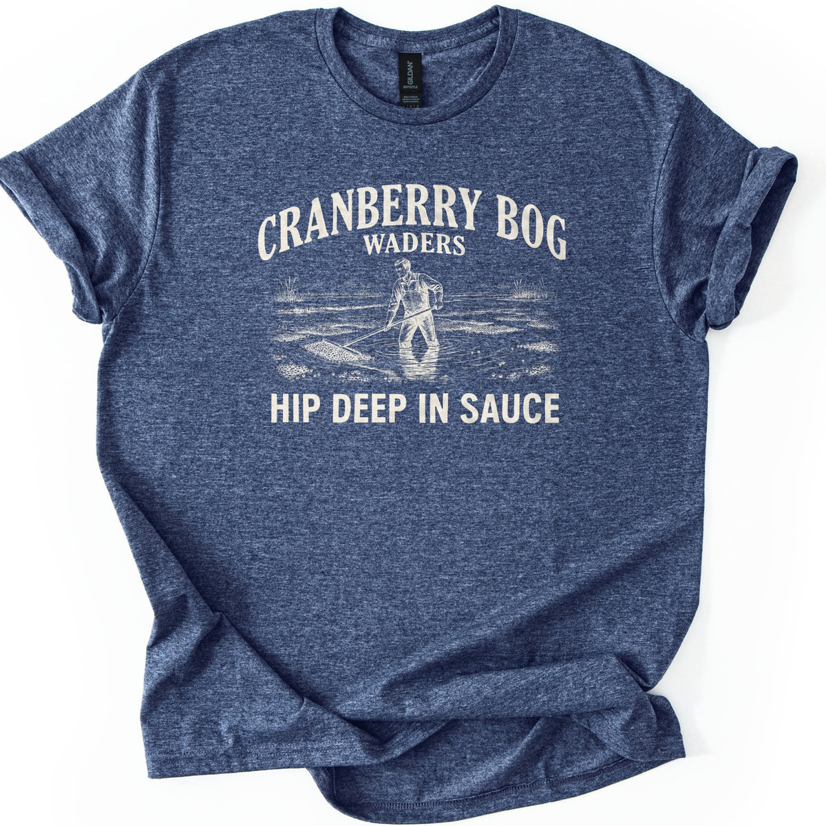 Cranberry Bog Waders Comfort Colors T-shirt in Heather Navy, laid flat. A cream etched drawing of a man in overalls standing hip deep in a flooded bog, raking floating cranberries, under the words Cranberry Bog Waders and above Hip Deep in Sauce. Sleeves cuffed, on white.
