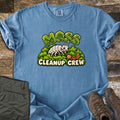 Moss Cleanup Crew Comfort Colors T-shirt in Blue Jean, laid flat. A smiling white cartoon bug sweeps a brown leaf with a broom atop green moss, below the word MOSS in mossy letters and above CLEANUP CREW. It lies on weathered wood with folded olive pants and brown hiking boots.