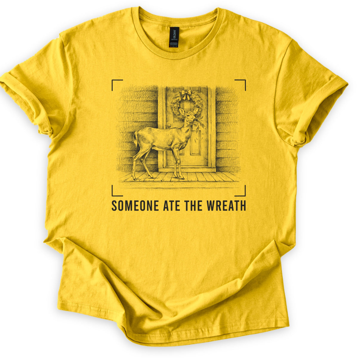 Someone Ate The Wreath Comfort Colors T-shirt in Daisy, laid flat. A pencil-style sketch of a deer on a wooden porch, a sprig of greenery in its mouth, below a bowed wreath on the door, framed by camera viewfinder corners above the words Someone Ate The Wreath.