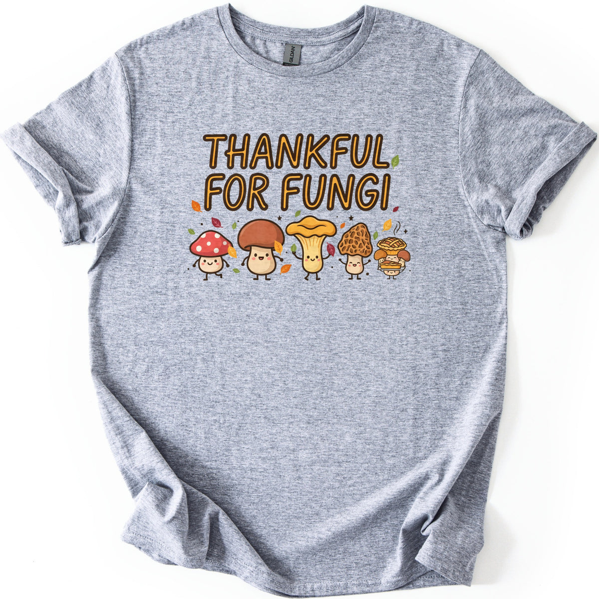 Thankful For Fungi Comfort Colors T-shirt in Sport Grey, laid flat. The words Thankful For Fungi in orange letters above five smiling cartoon mushrooms, one holding a pie, with autumn leaves around them. The shirt has rolled sleeves on a white background.