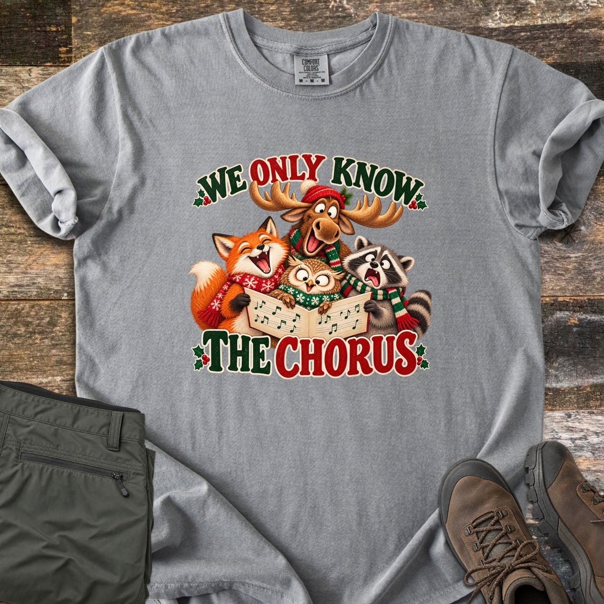 We Only Know the Chorus Comfort Colors T-shirt in Grey, laid flat. A cartoon moose in a red hat, a fox, an owl and a raccoon in scarves sing from a songbook between the words We Only Know The Chorus. Shown on wood boards with green pants and brown boots.